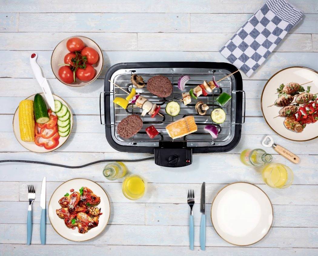 A tabletop grill on a wooden table with vegetables, meat, and skewers. Surrounded by plates, tomatoes, glasses of drinks, and cutlery.