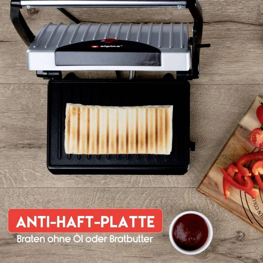 A sandwich is being grilled in a panini press. Bottom left reads 'NON-STICK PLATE - Grill without oil or cooking butter'. To the right, a wooden board with sliced tomatoes lies.