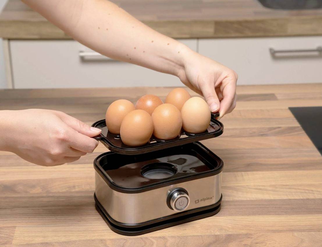 A person places a tray with six eggs onto an egg boiler in a kitchen. The egg boiler is situated on a wooden worktop.
