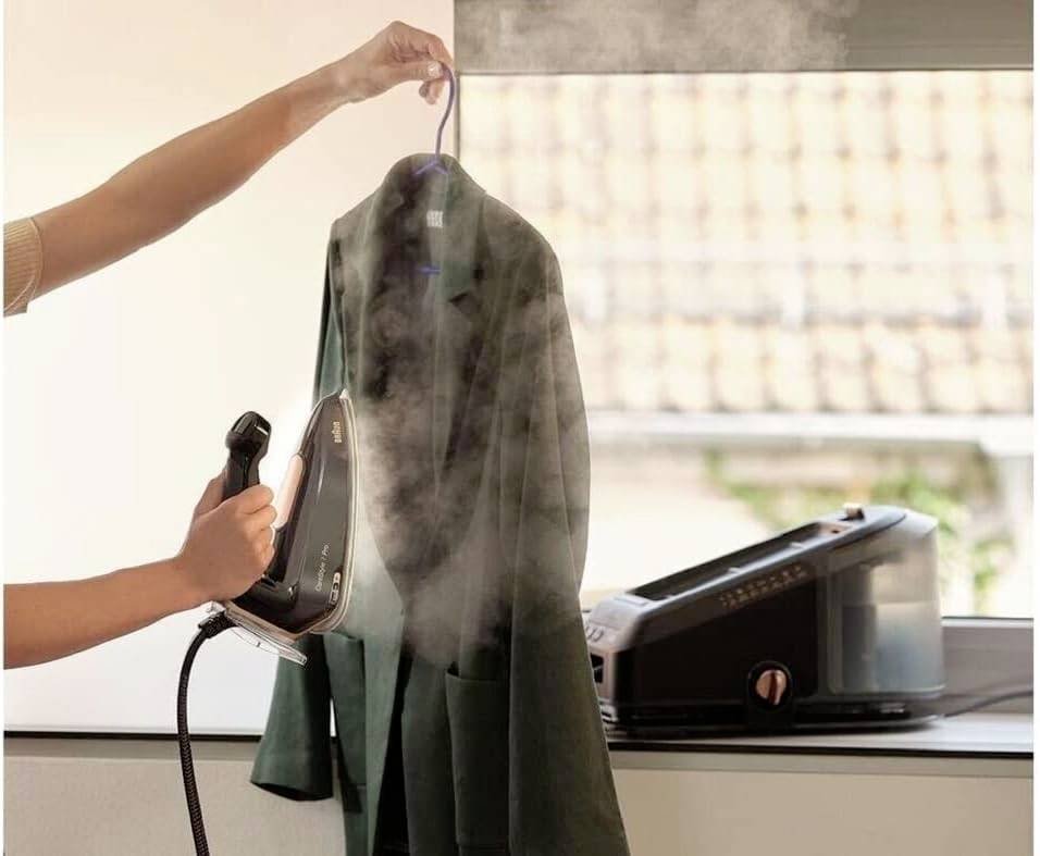 A person is steam ironing a suit jacket hanging on a clothes hanger. In the background, a steam ironing station sits on the windowsill.