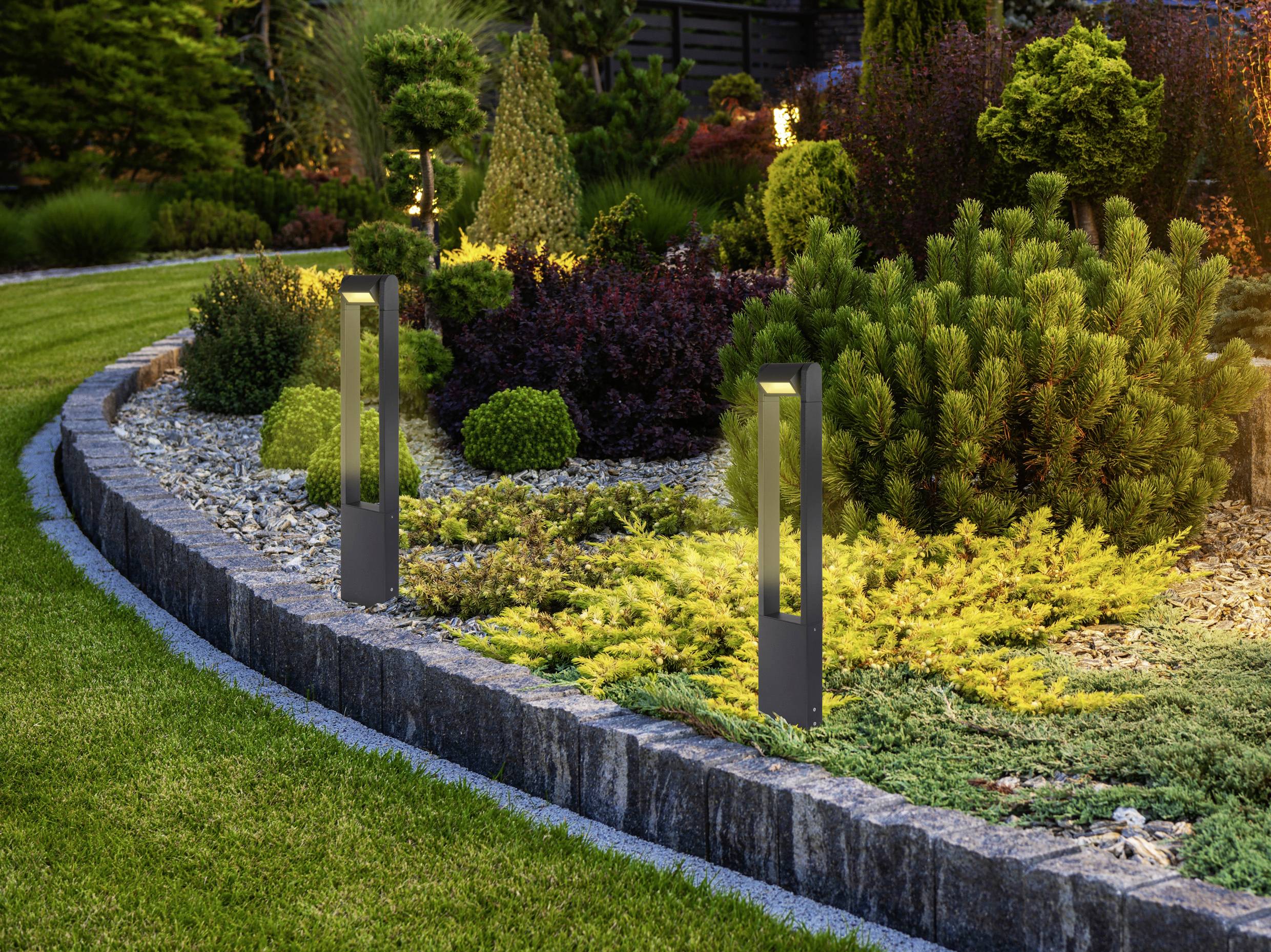 A landscaped garden with neatly trimmed shrubs and decorative lighting fixtures along a stone border, surrounded by green grass.