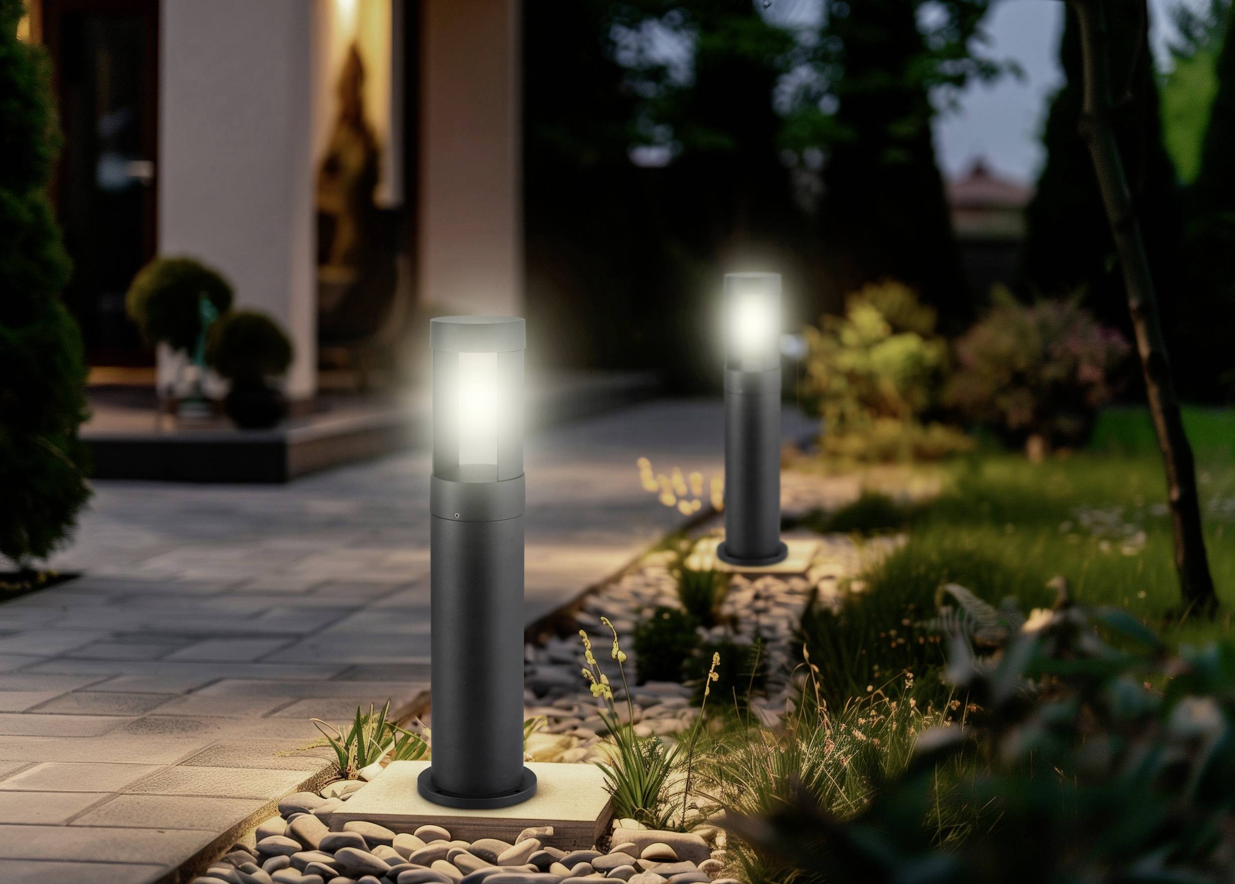 Two modern outdoor lamps illuminate a path lined with stones and grass, leading to a house with a warmly lit interior.