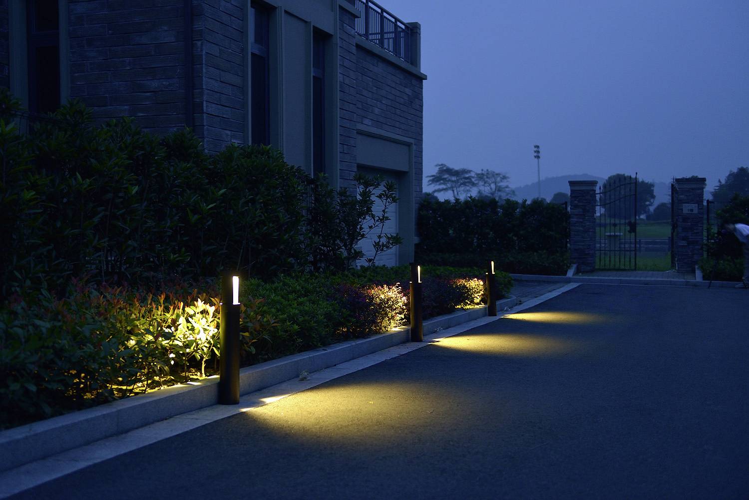 A dimly lit walkway at dusk, lined with modern path lights and greenery, leading past a building towards a gated entrance in the background.