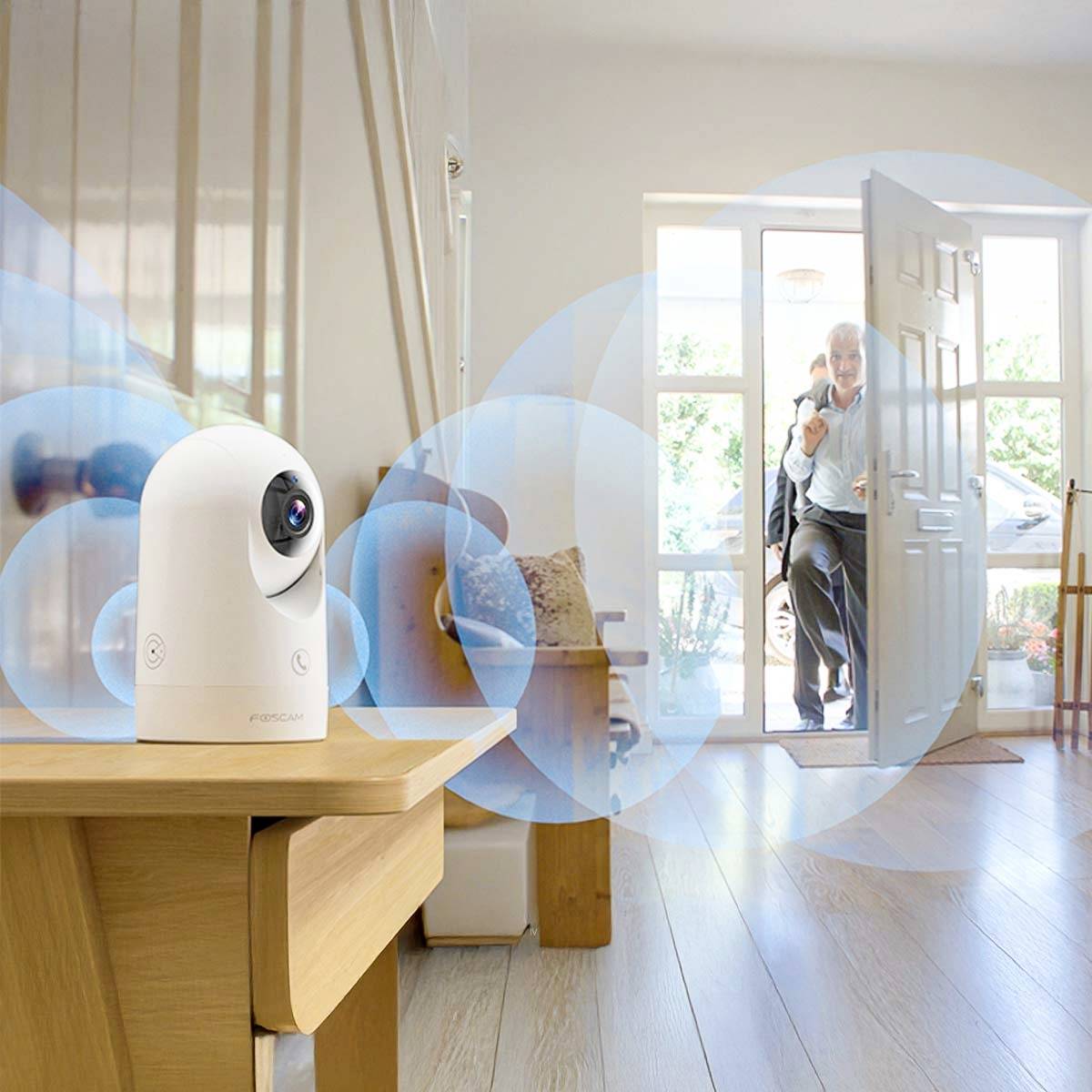 A white indoor camera on a table, monitoring a man entering a house through an open door.