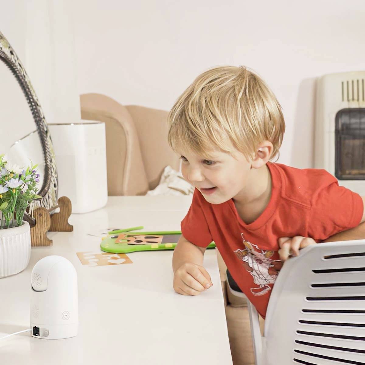 A small boy curiously observes a baby monitor on a table, surrounded by plants and everyday objects.
