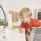 A small boy curiously observes a baby monitor on a table, surrounded by plants and everyday objects.