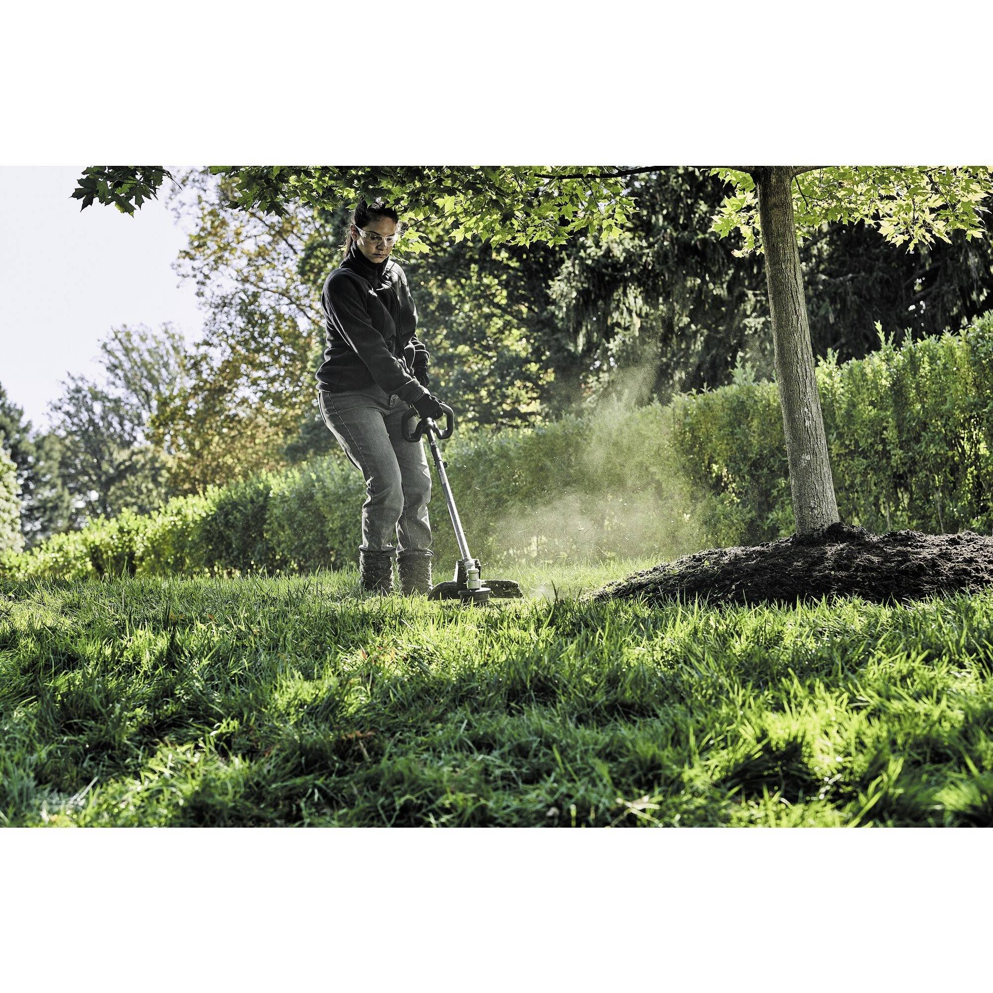 A person is using a lawn strimmer to cut grass beneath a tree in a sun-drenched garden.