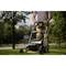 A person is mowing a green lawn with a lawnmower in daylight. Blurry trees can be seen in the background.