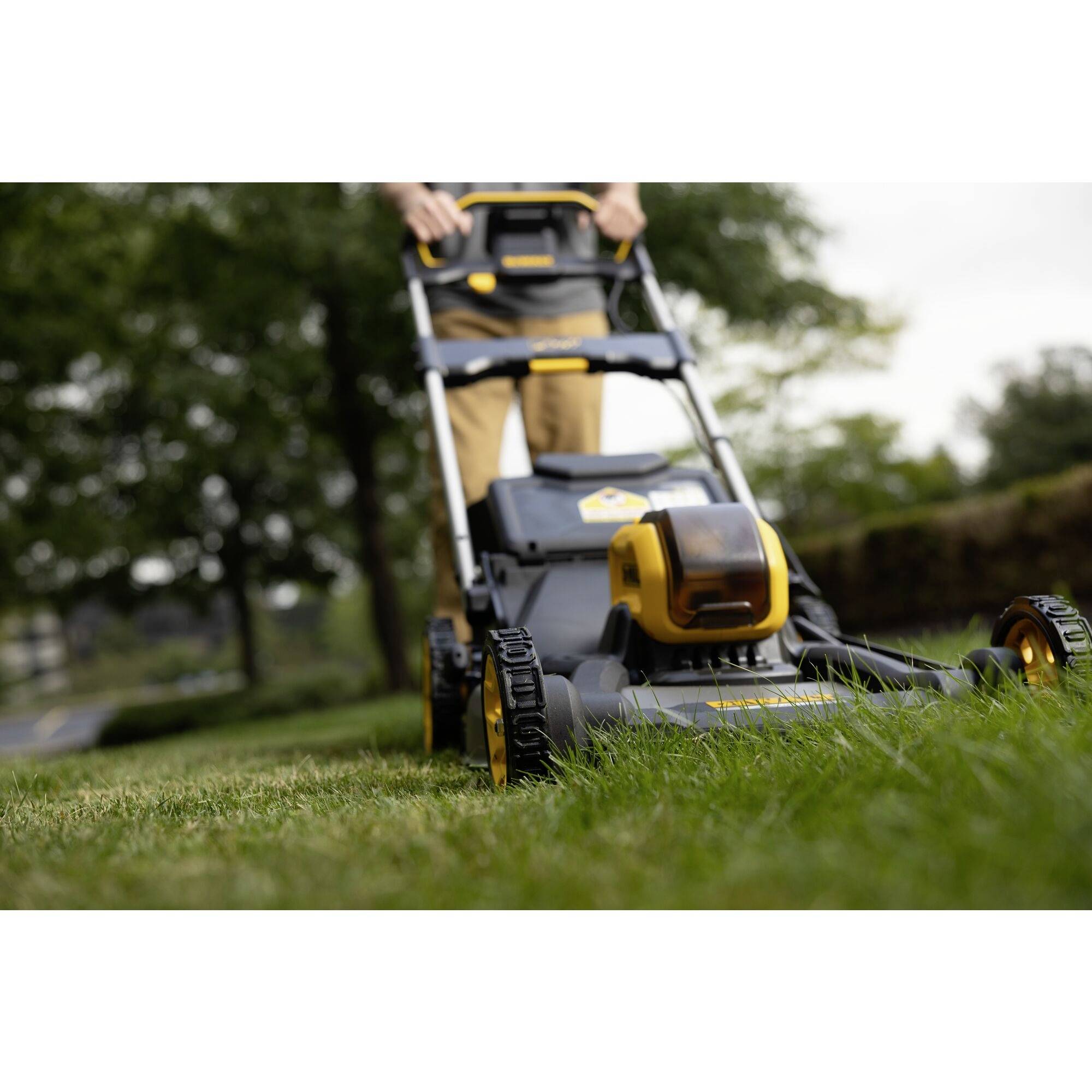 A person is mowing the lawn with a lawnmower on a green lawn. Trees and bushes can be seen in the background.