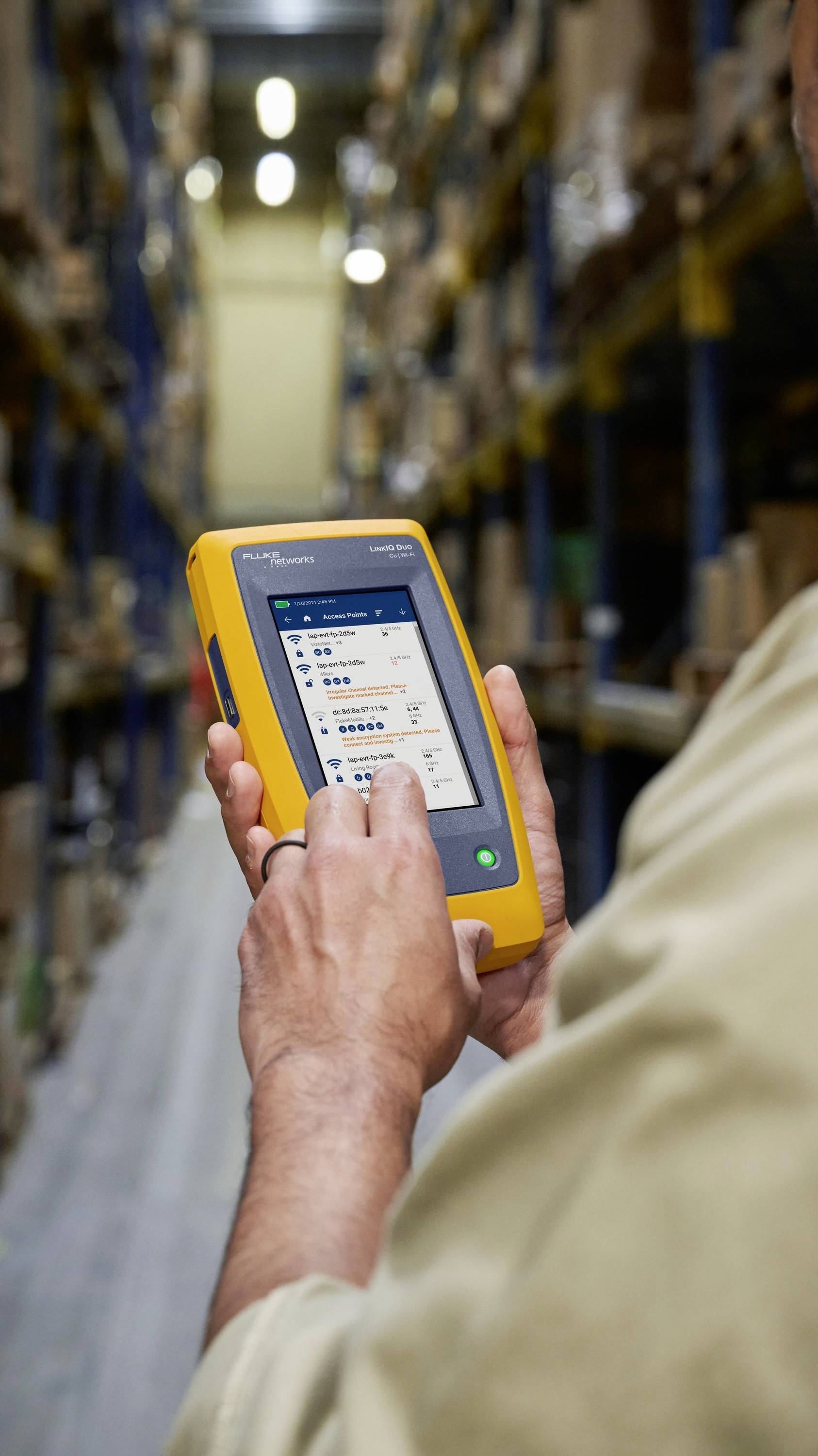 A person in a warehouse is operating a handheld touchscreen device for data capture. Shelves with crates are visible in the background.