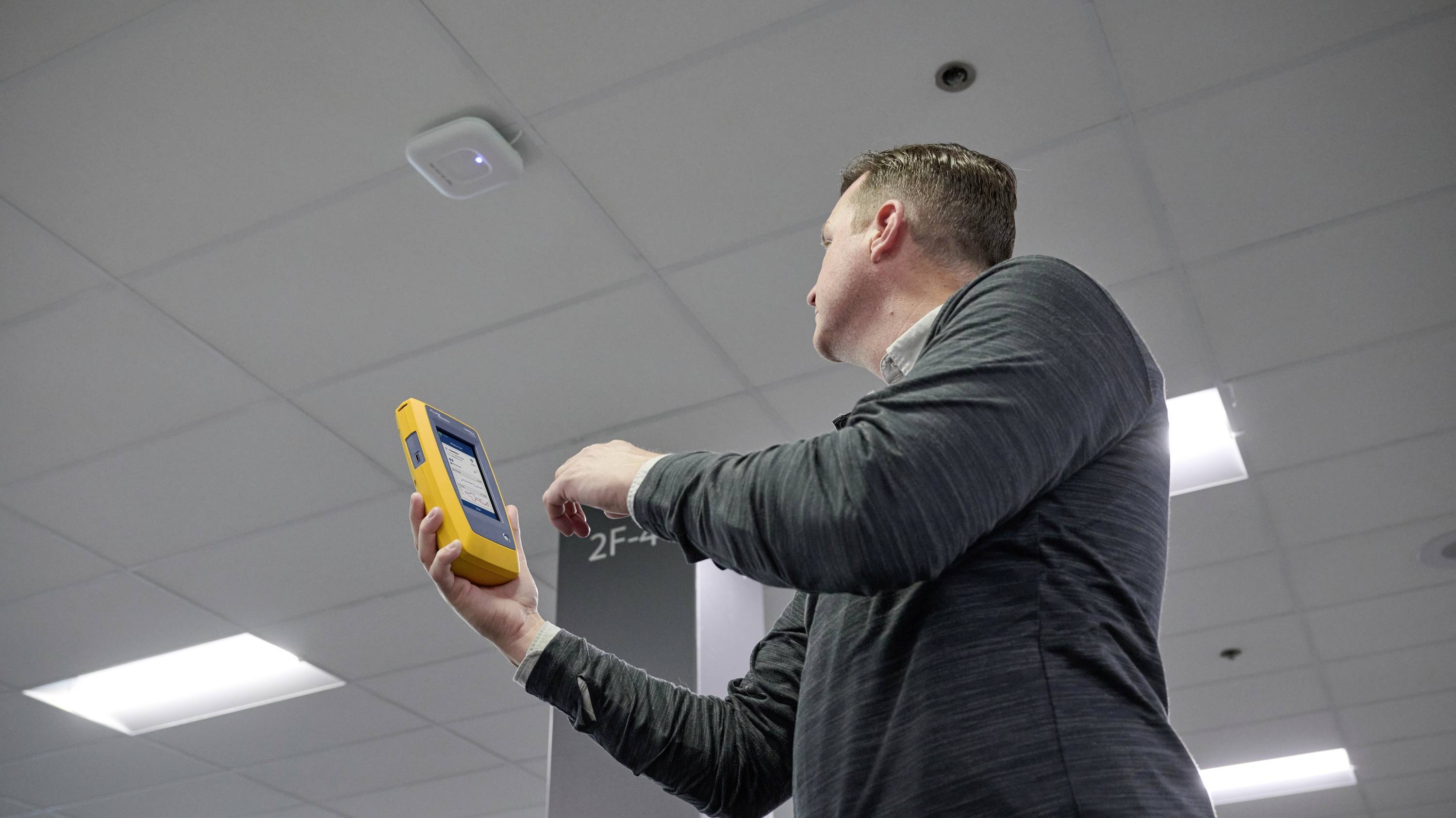 A man is holding an electronic device in a room with a suspended ceiling. The device appears to be used for measurement or analysis.