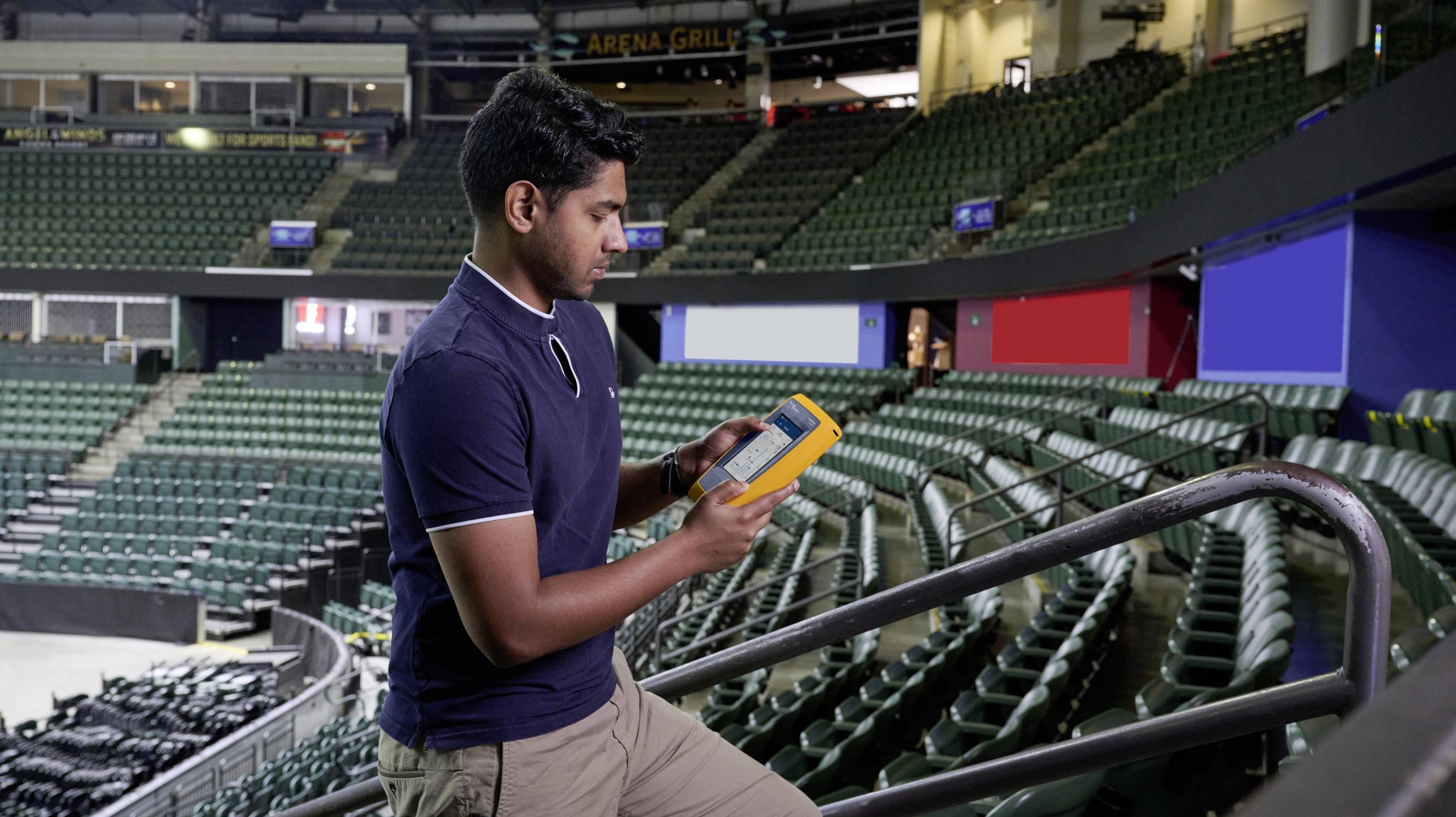 A man stands in an empty arena, looking down at an electronic device. The rows of seats are vacant, and the lighting is dimmed.