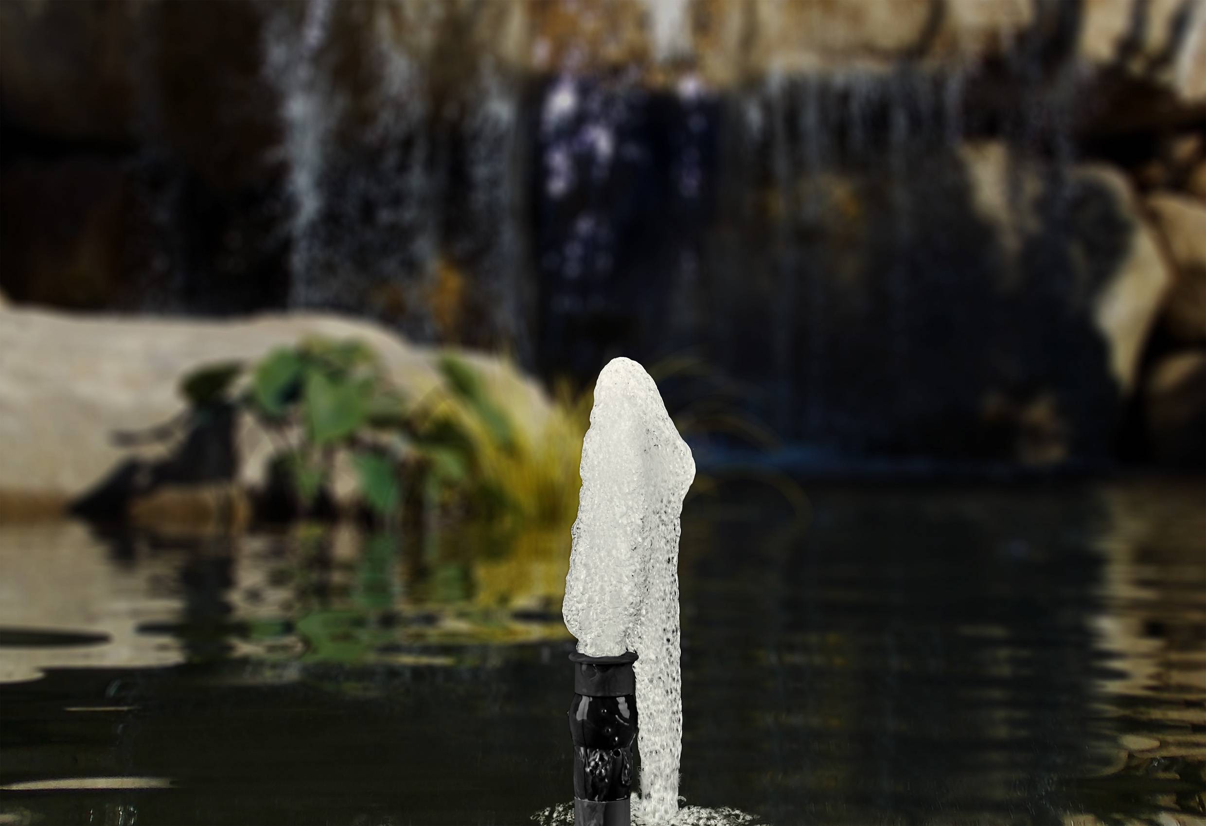 A small fountain in the foreground of a pond, with a splashing waterfall and rocks in the background.