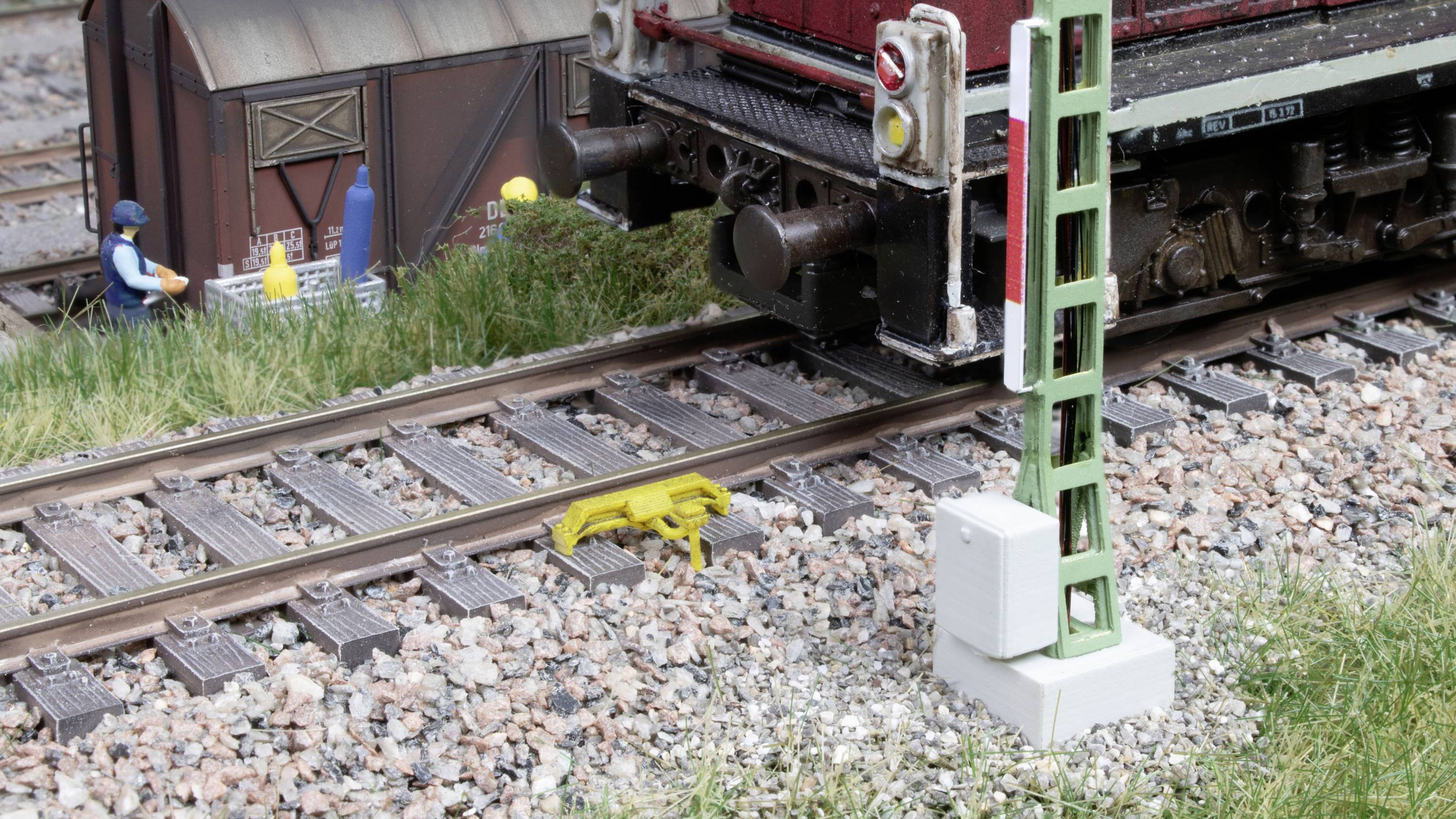 A model railway with tracks, a train, and a yellow track barrier. In the background, a model building and a figure.