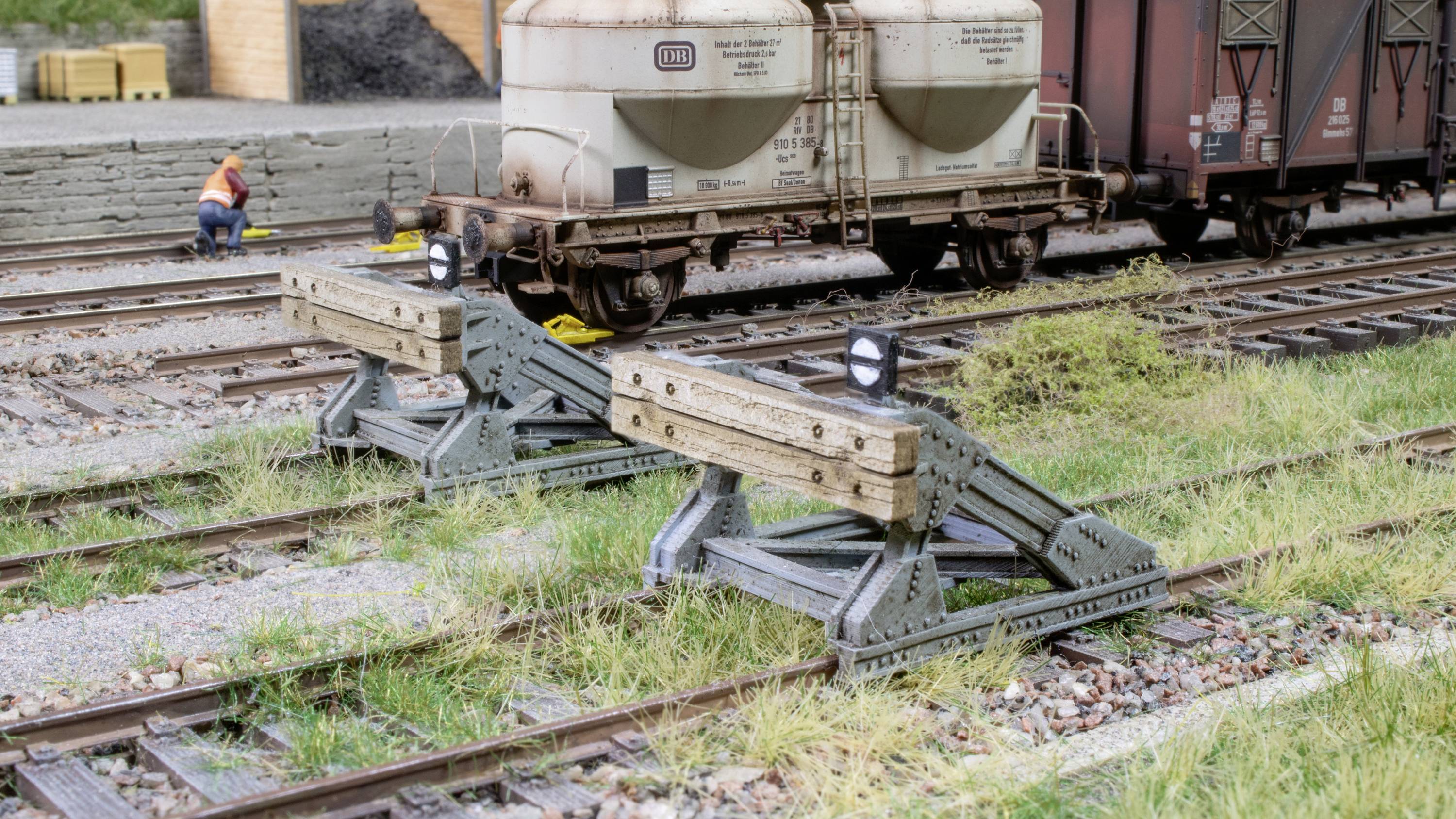 Two buffer stops halt rails on a railway site, with goods wagons standing behind. A worker in an orange high-visibility vest is in the background.