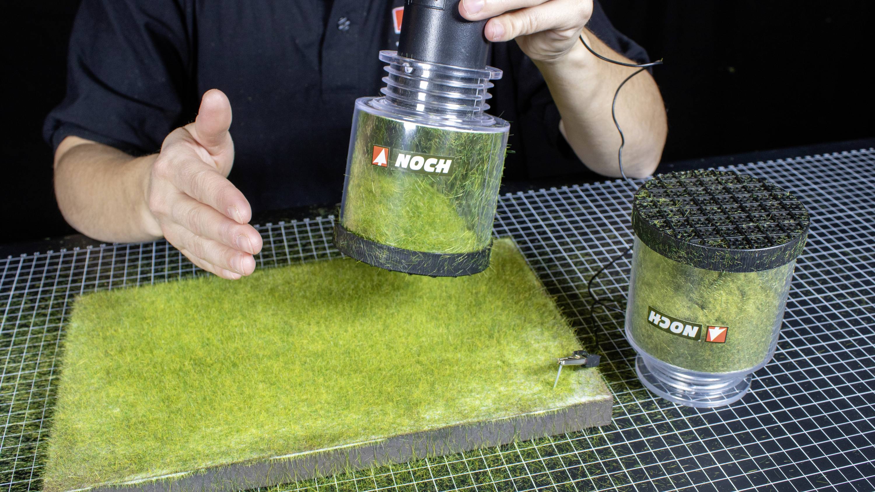A person demonstrates the use of a grass applicator on a model building plate. Two devices with grass fibres are visible.