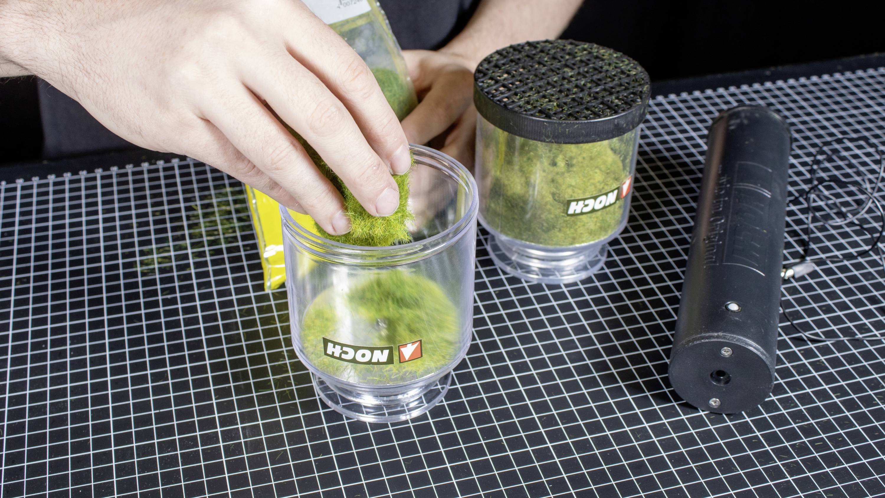 A hand fills green granules into a transparent container with a Nock logo. Another container and a black device are beside it.