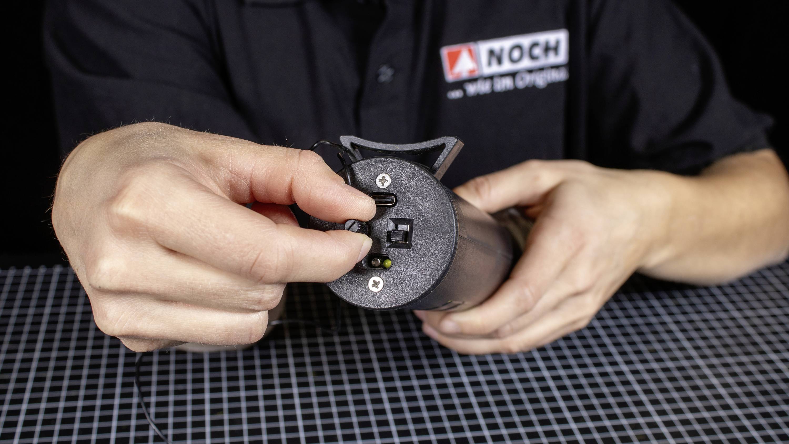 A person is holding a small electrical device with connection ports. A logo is visible on the person's shirt.