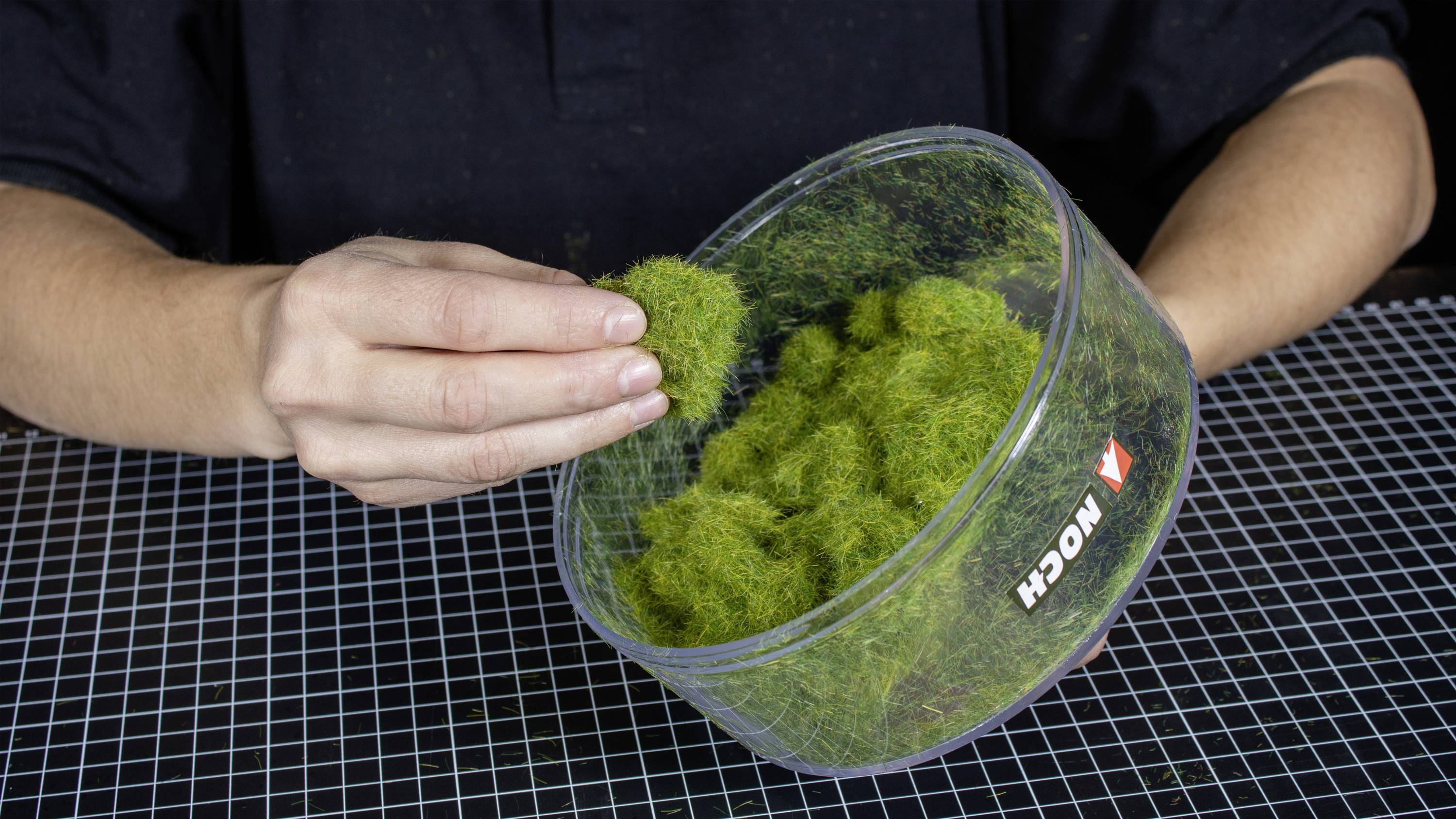 A person is holding a transparent bowl filled with green moss substitute, resting on a chequered surface.