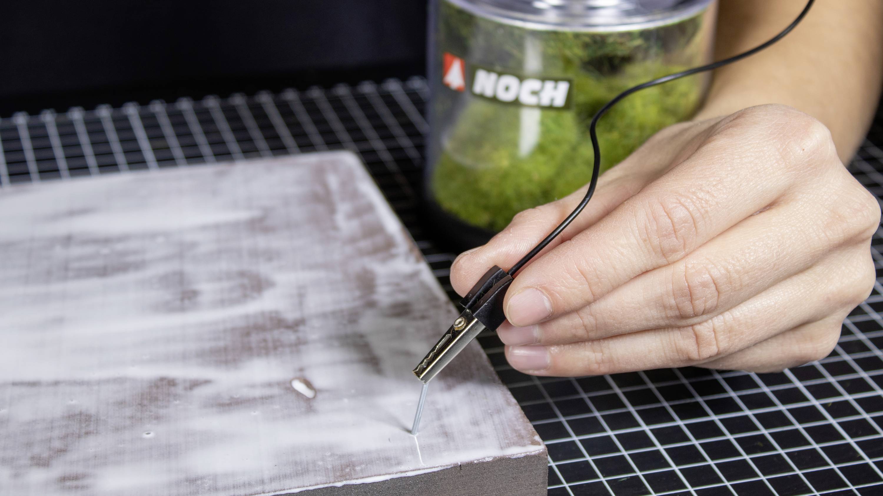 A hand is working on a rectangular piece of modelling material with a fine tool. In the background, a container with the 'Noch' logo.