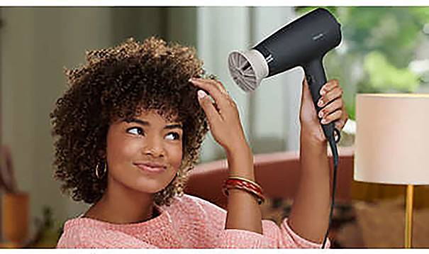A person is drying their curly hair with a hairdryer indoors. She is wearing a pink top and has a slight smile.