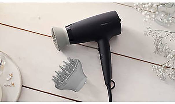 A black hairdryer with a diffuser attachment lies on a wooden table. Next to it is another attachment, surrounded by decorative branches.