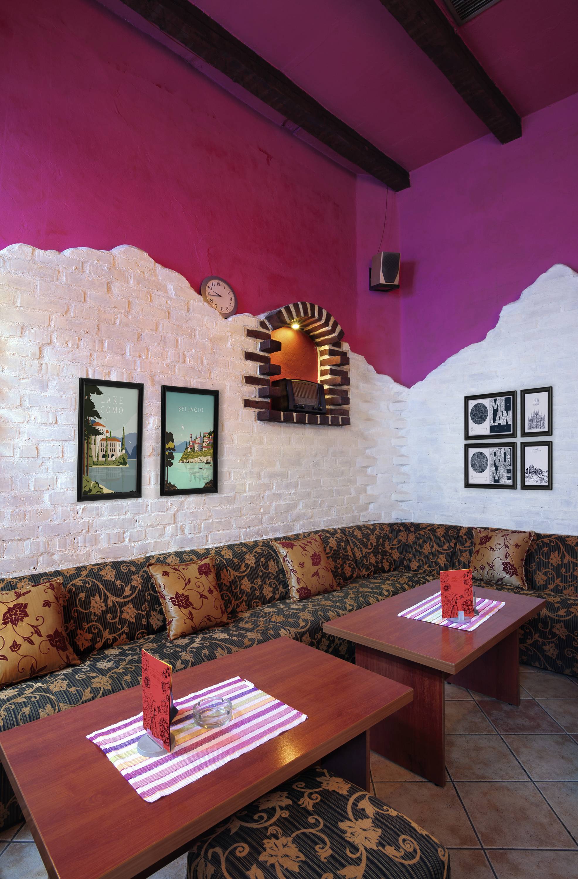 Cosy café with white and pink walls, dark wooden beams and patterned sofas around wooden tables. Colourful posters and photographs adorn the walls.