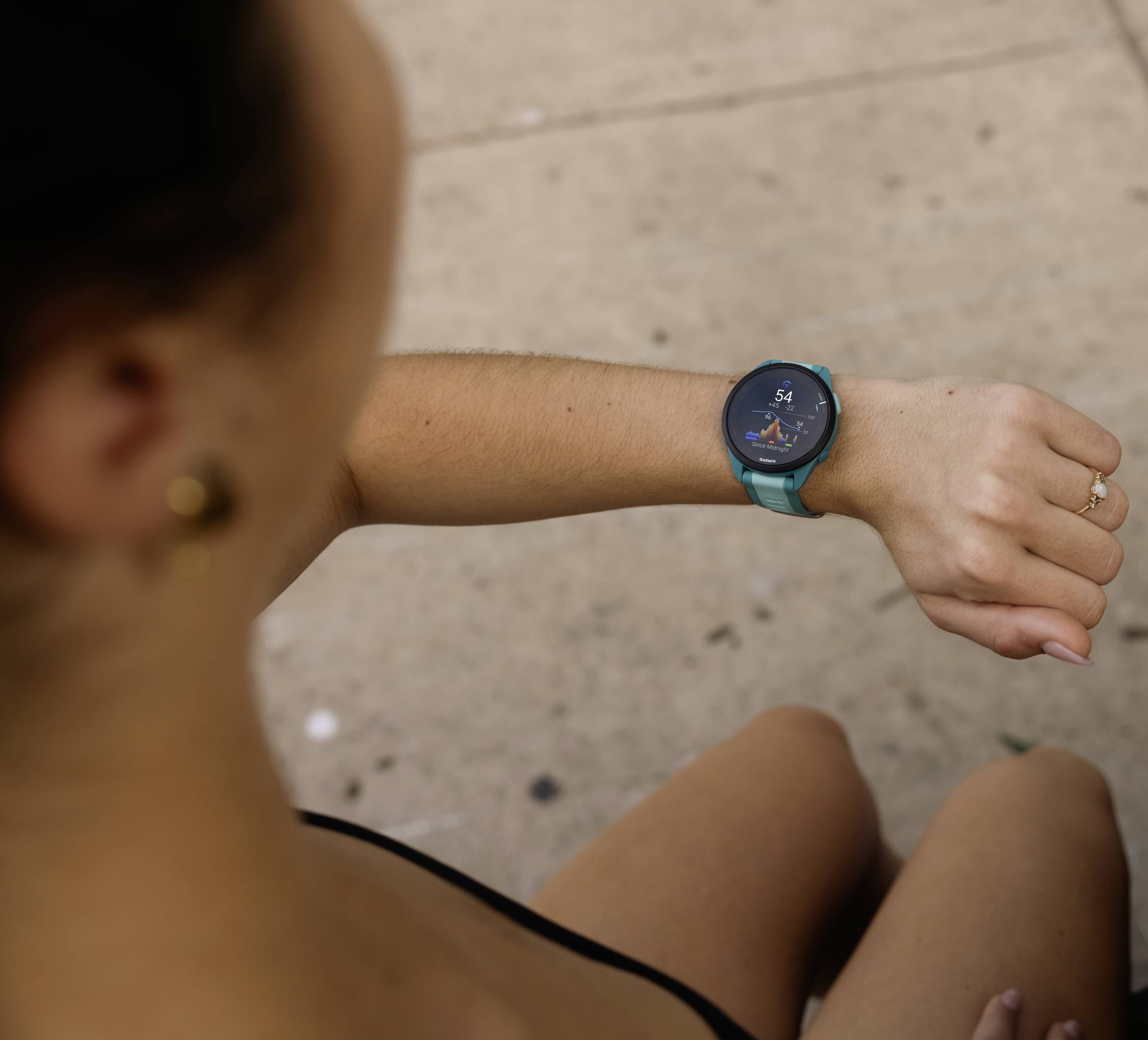 A person looks down at a smartwatch on their wrist, which displays information. They are sitting on the ground next to a pavement.