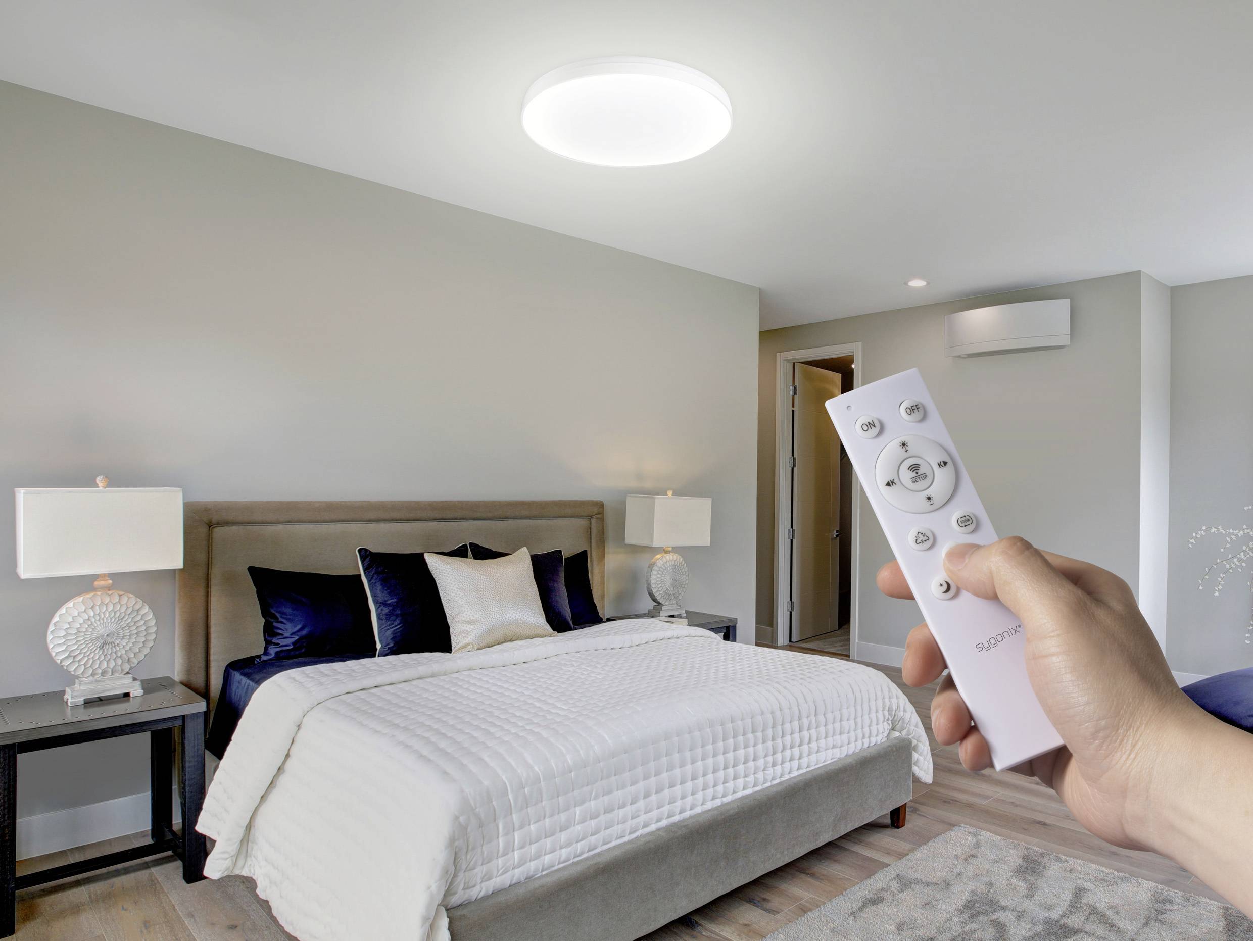 A modern bedroom with a bed, decorative pillows, and side lamps. A hand holds a remote towards an illuminated ceiling light.