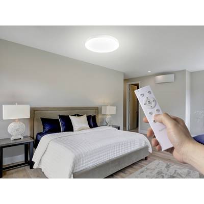A modern bedroom with a bed, decorative pillows, and side lamps. A hand holds a remote towards an illuminated ceiling light.