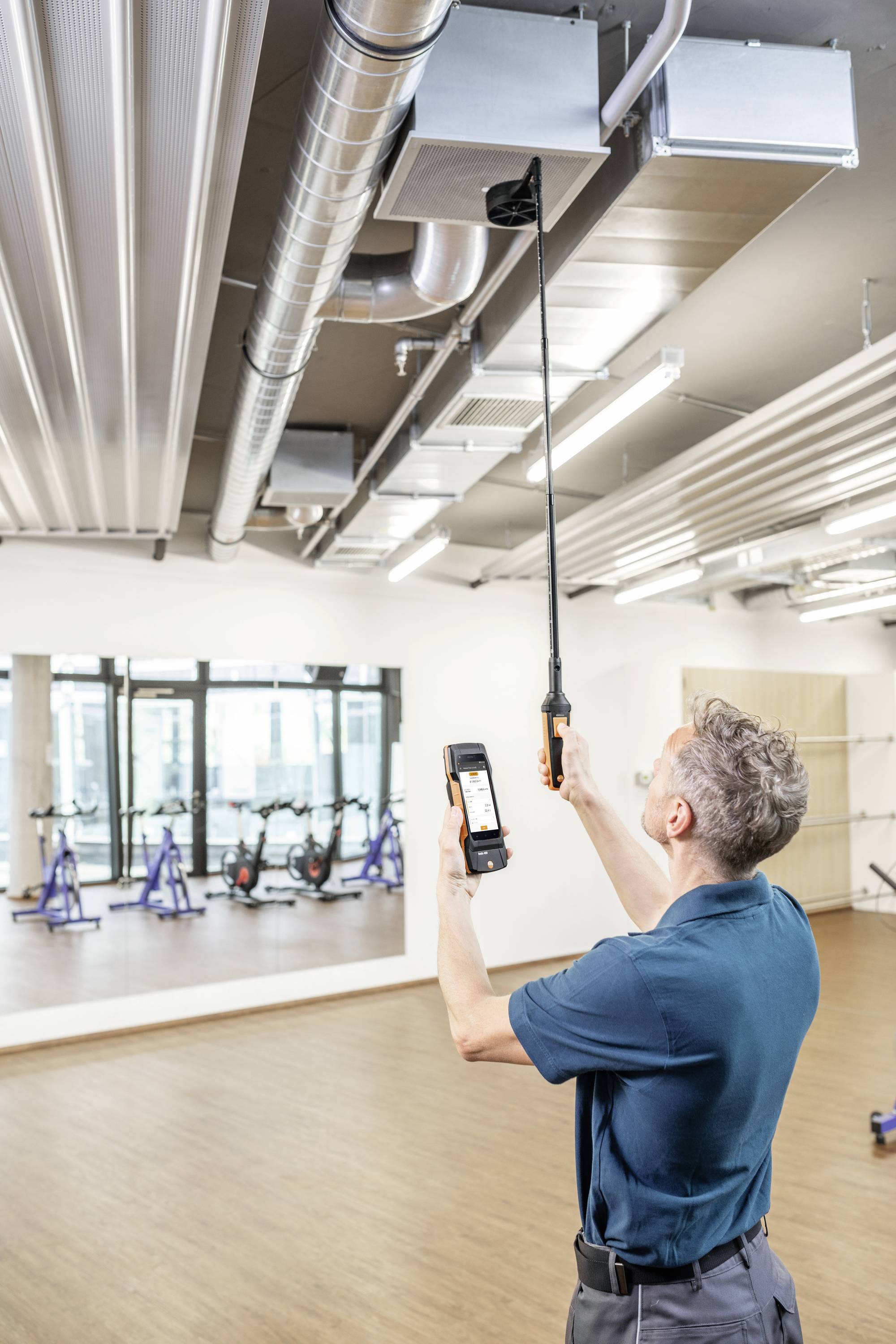A man is checking the ventilation system with a specialist measuring device in a room with a wooden floor and stationary bicycles.