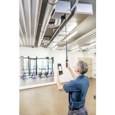 A man is checking the ventilation system with a specialist measuring device in a room with a wooden floor and stationary bicycles.