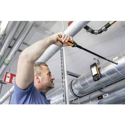 A man is inspecting a ventilation system with a long measuring device. A digital display shows results. The environment is industrial.