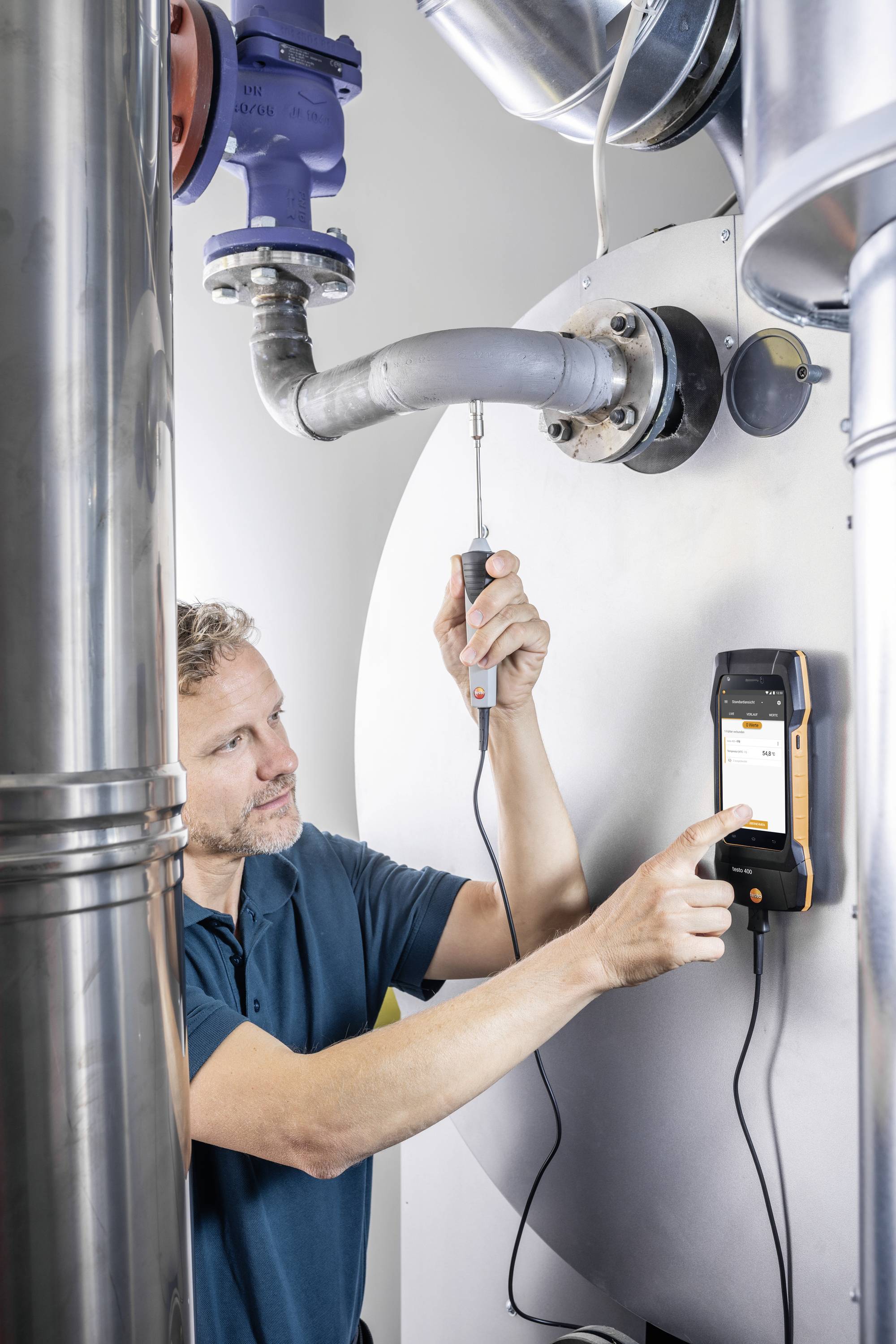 A man is using a measuring device to check heating pipes in a boiler room. He is holding a sensor against the pipe.
