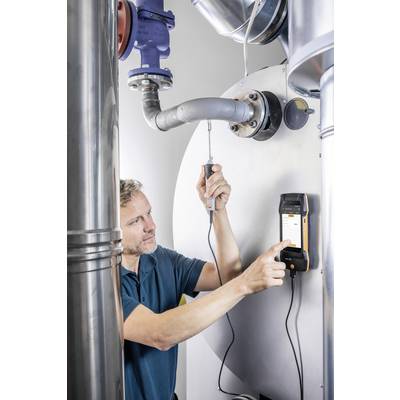 A man is using a measuring device to check heating pipes in a boiler room. He is holding a sensor against the pipe.