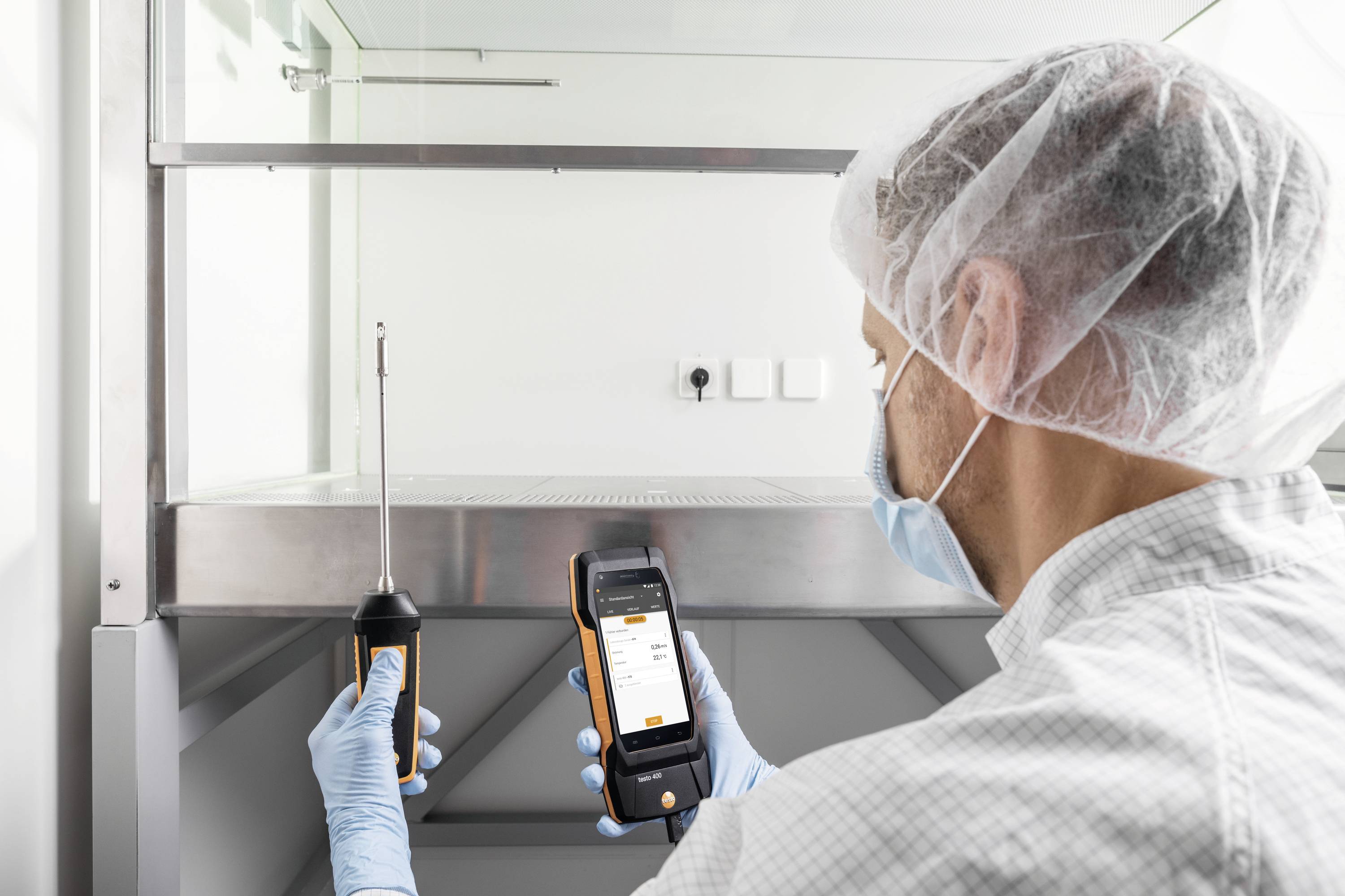 A technician is measuring air quality in a laboratory using a mobile device. He is wearing protective clothing and a hair net.