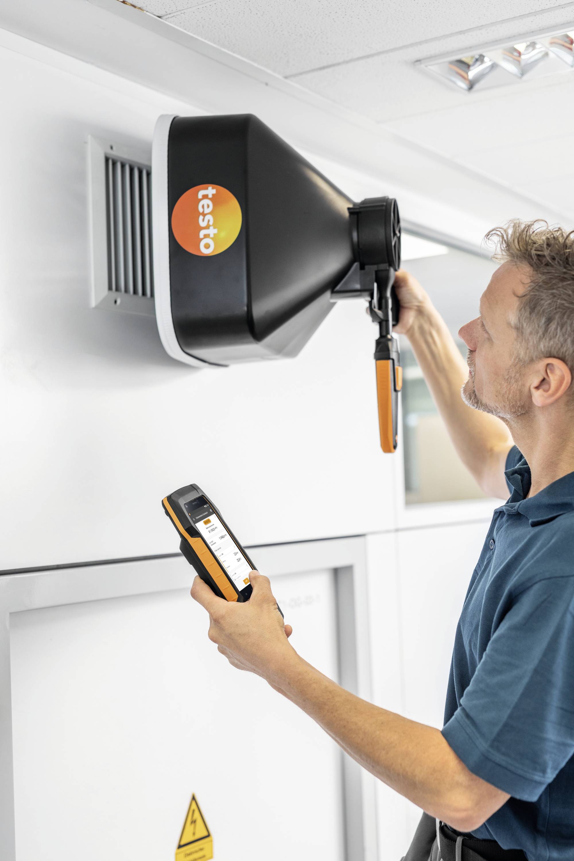 A man is measuring the airflow at a ventilation opening in a room using a device.