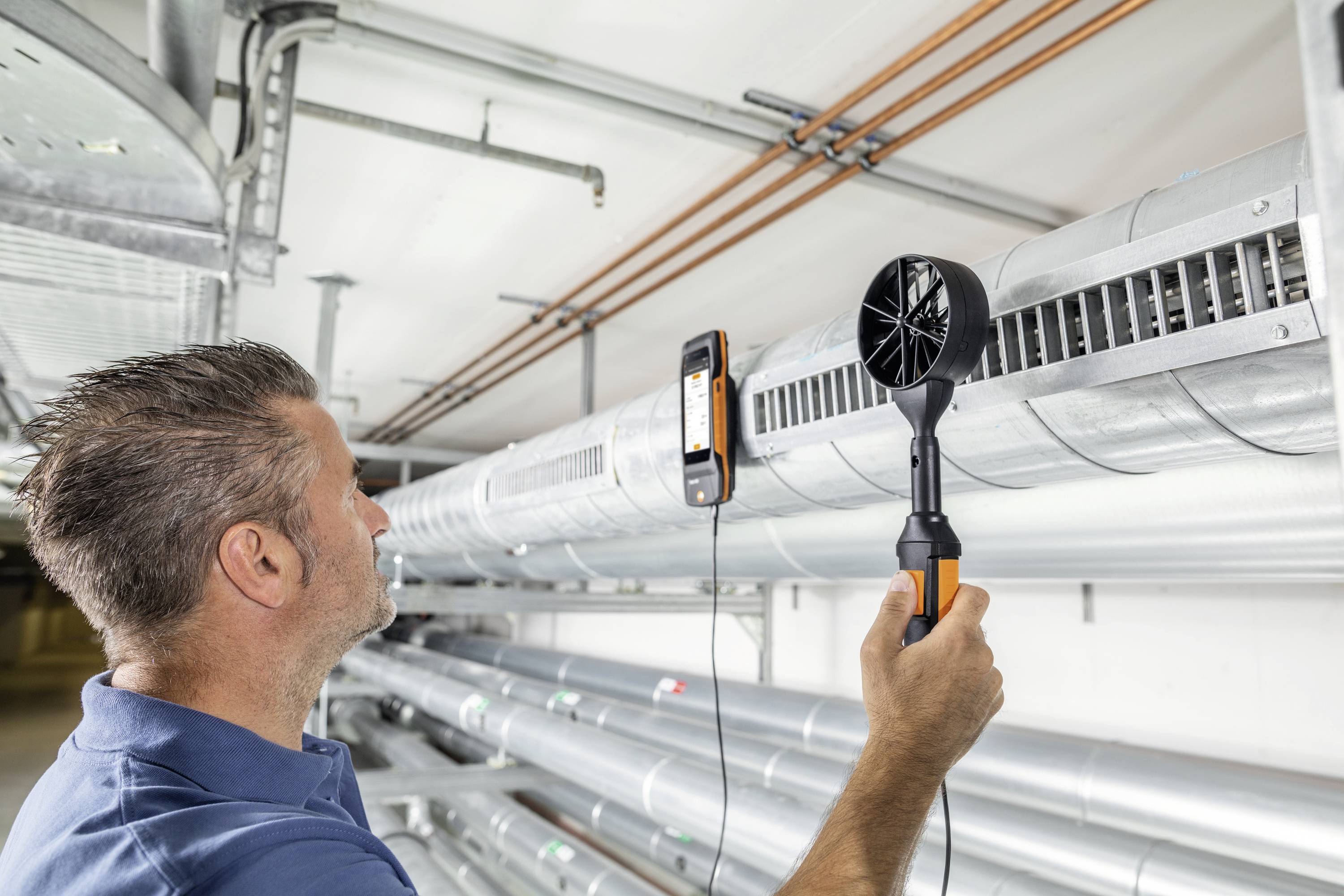 A technician is measuring airflow in a large ventilation duct with an anemometer in an industrial setting.