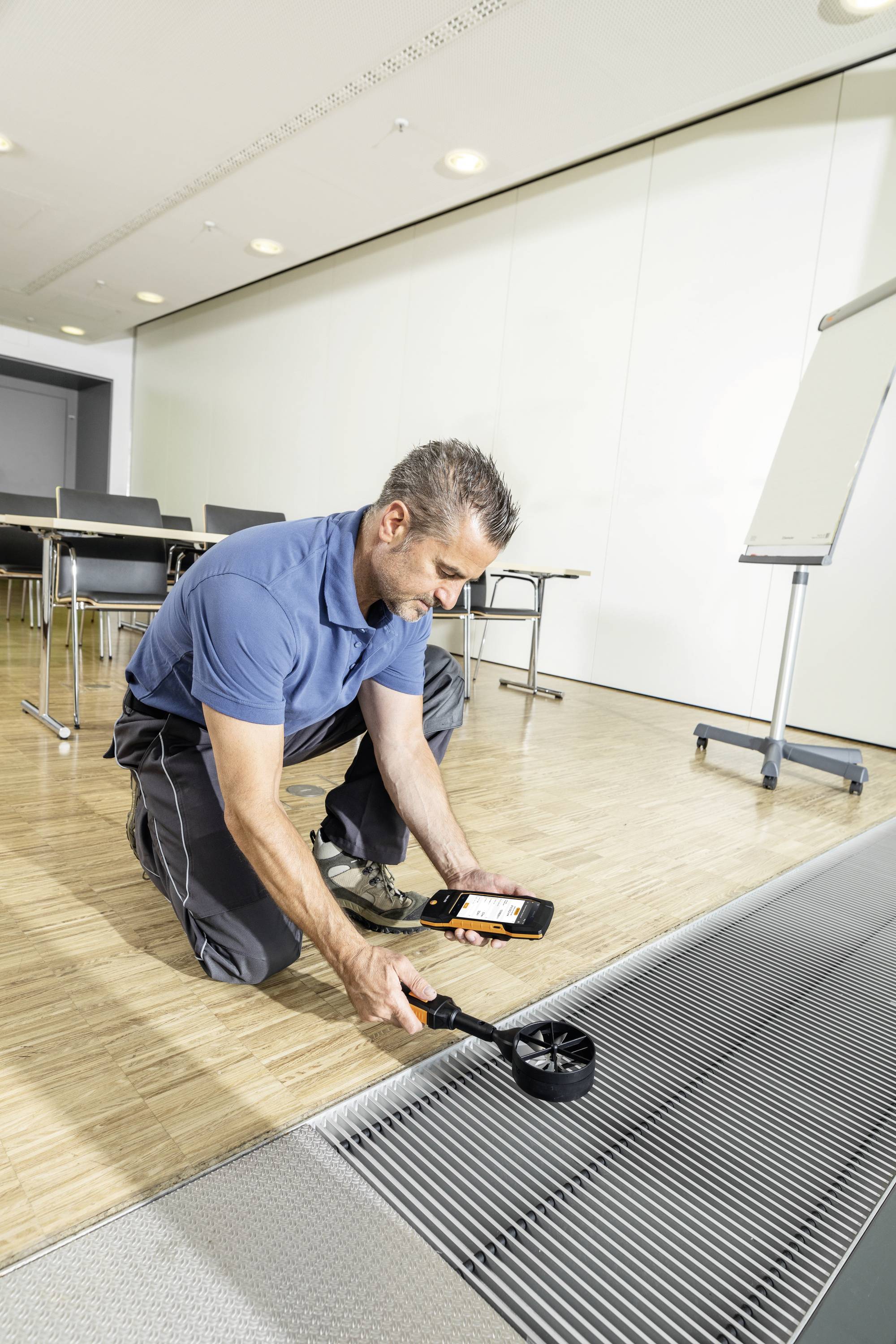 A man in work attire is measuring the airflow at a ventilation grille in a modern office using an anemometer.
