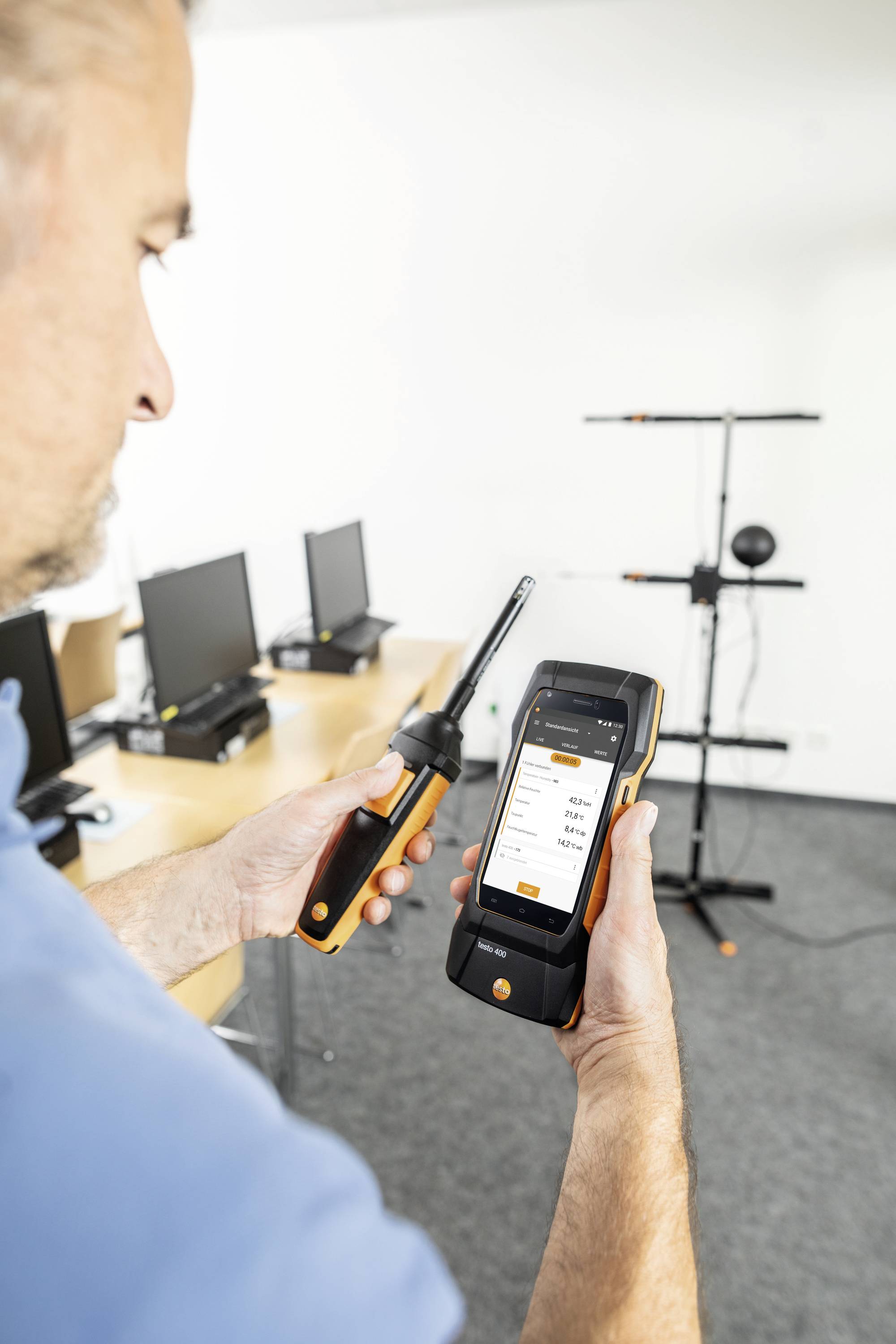 A person is holding a measuring device in an office. The screen of the device displays measurement data. Computers are visible on desks in the background.