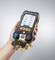 A hand is holding a digital pressure gauge with a display screen showing gas pressure values. The device has multiple valves and is designed for HVAC applications.