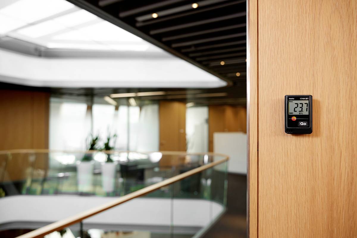 A digital thermometer shows 23.7 degrees Celsius in a modern office building with glass balustrades and plants in the background.