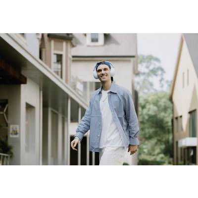 A smiling man with headphones is walking past modern buildings. He is wearing a blue shirt and white trousers. Mood: relaxed.