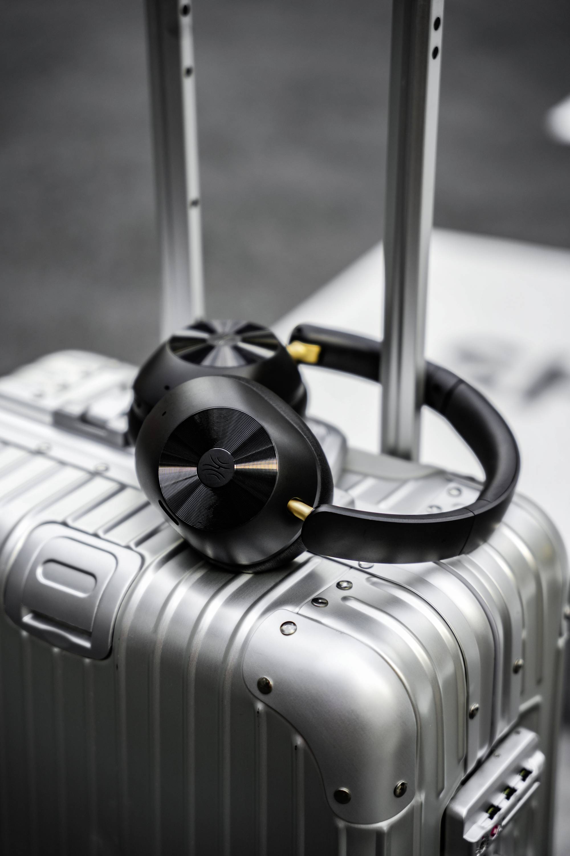 Black headphones lie on a silver wheeled suitcase. The environment appears like an airport or train station, suggesting readiness for travel.