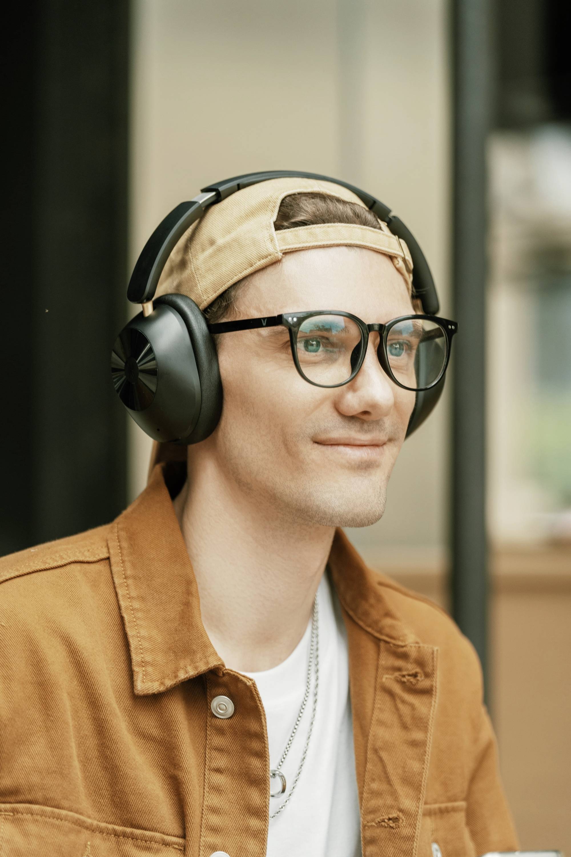 A person wearing glasses and headphones, dressed in a light shirt and brown jacket, sits smiling and looking to the side.
