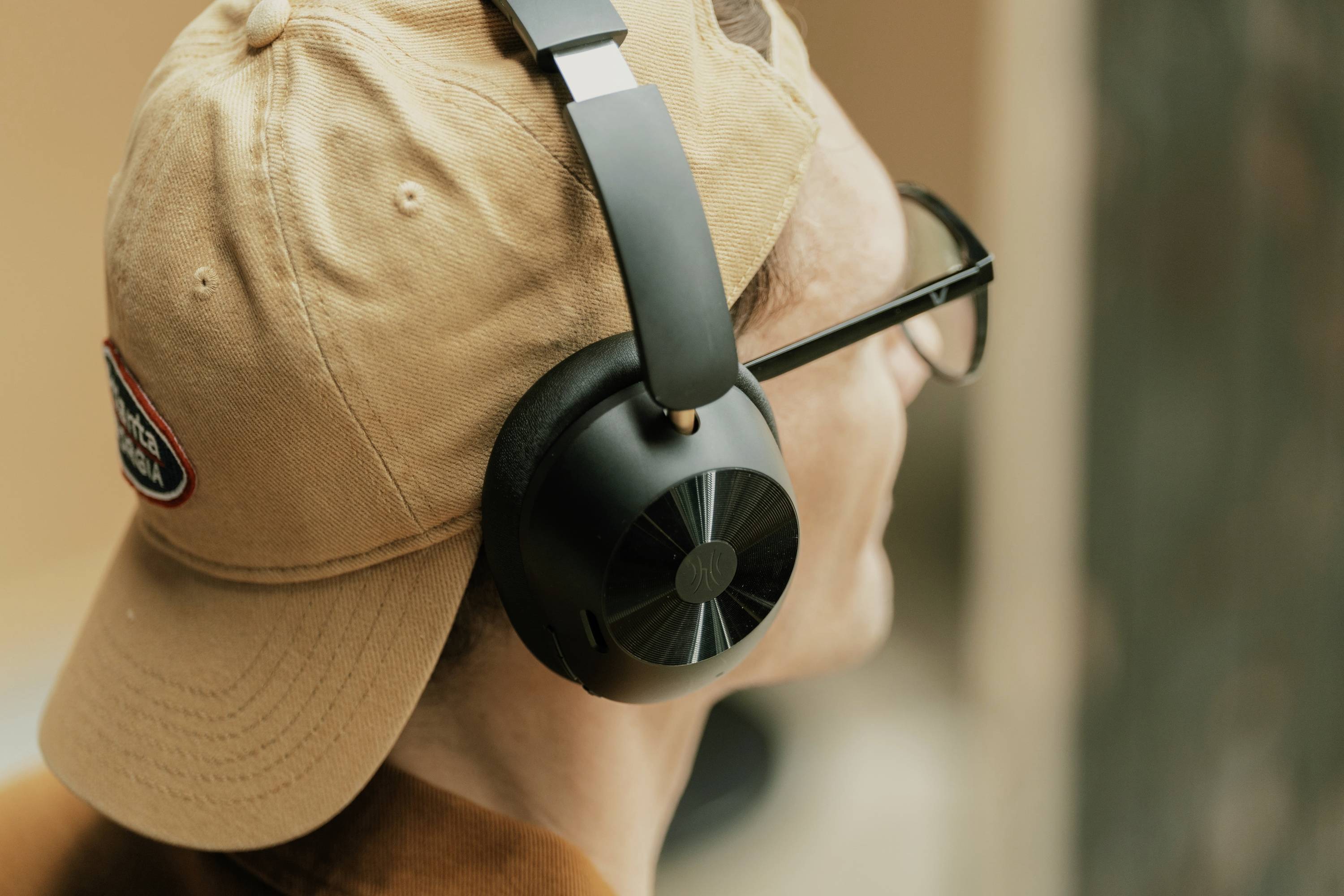 A person wearing glasses and a baseball cap has black headphones on and is looking into the distance. The background is out of focus.