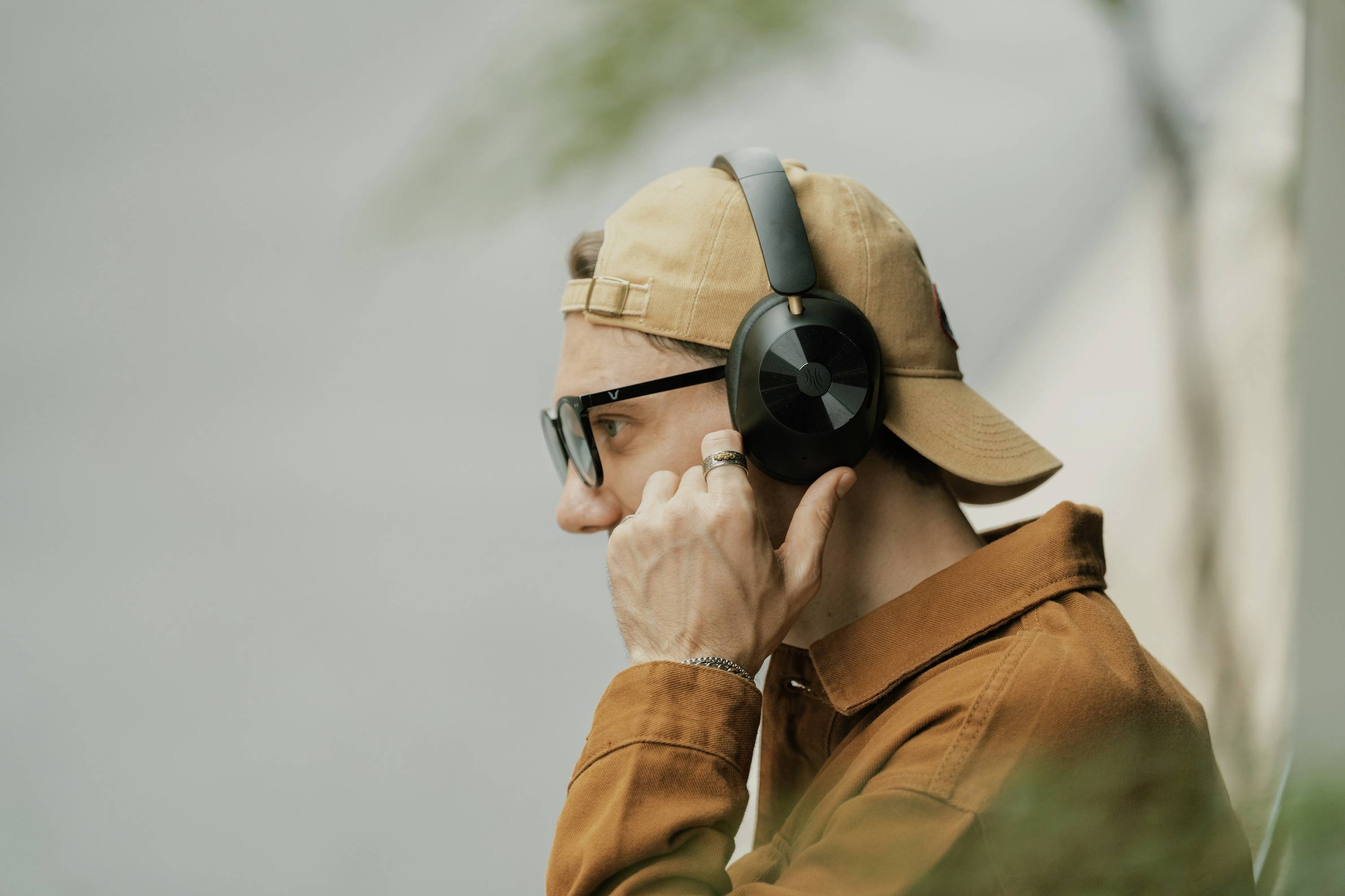 A man wearing a brown cap and headphones is listening to music. He is wearing glasses and is seen from the side, against a blurred background.