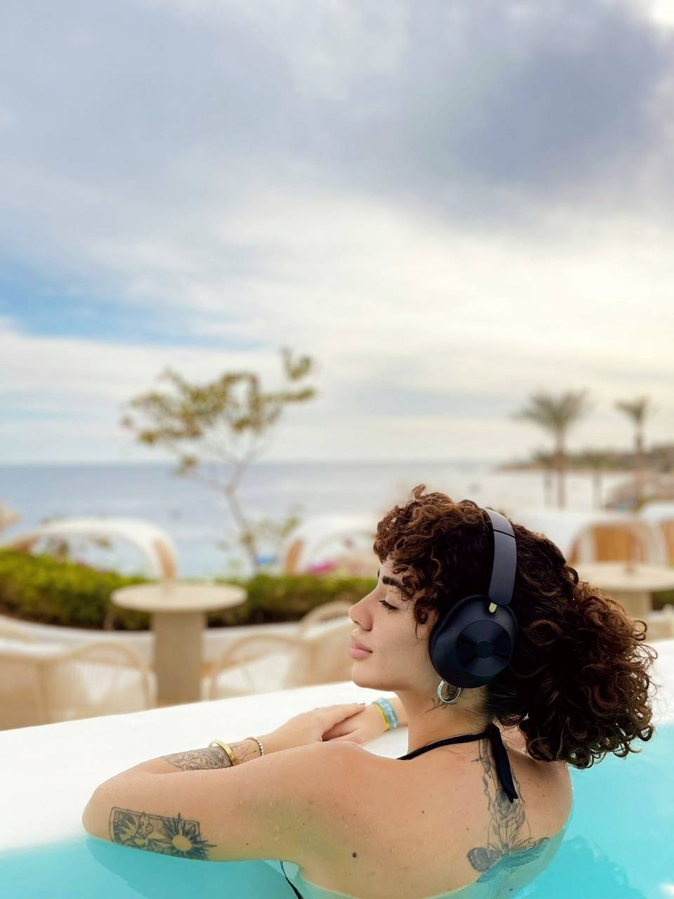 A person is relaxing in a pool, looking out to sea and wearing headphones. In the background, sky, trees and sunloungers can be seen.