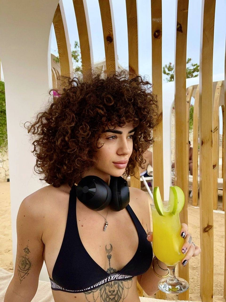 A person wearing headphones is sitting on a beach, holding a yellow drink with a lime slice. Parasols are visible in the background.