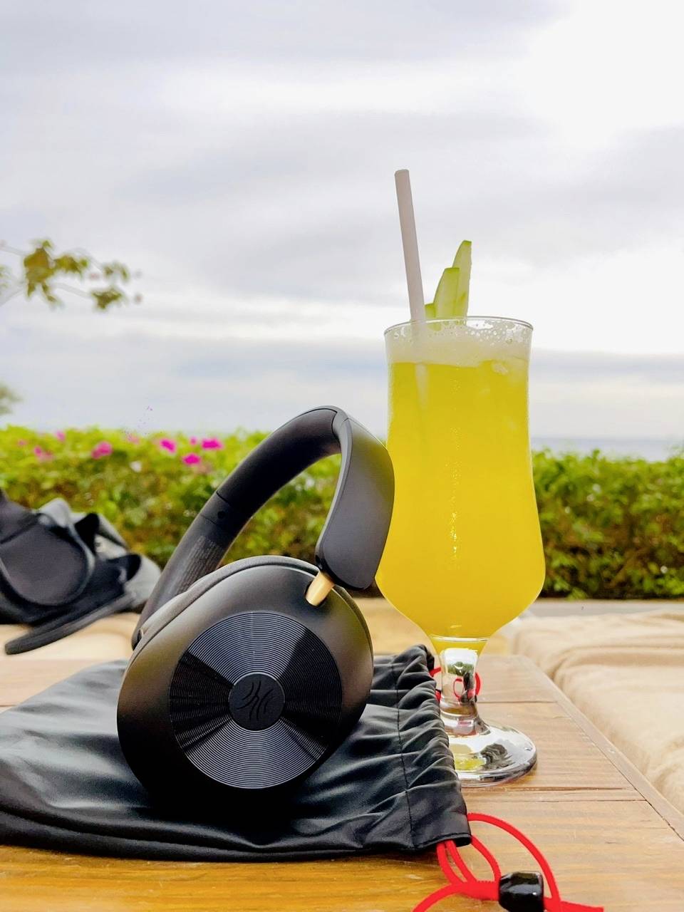 A black pair of headphones lies on a table next to a large glass with a yellow drink and a slice of cucumber. The background is a cloudy sky.
